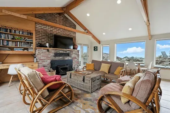 - Cozy living room with vaulted ceilings, rattan furniture, and vibrant pillows  - Brick fireplace with mounted TV for a warm and inviting atmosphere  - Large windows offer sweeping views of the dunes and ocean beyond