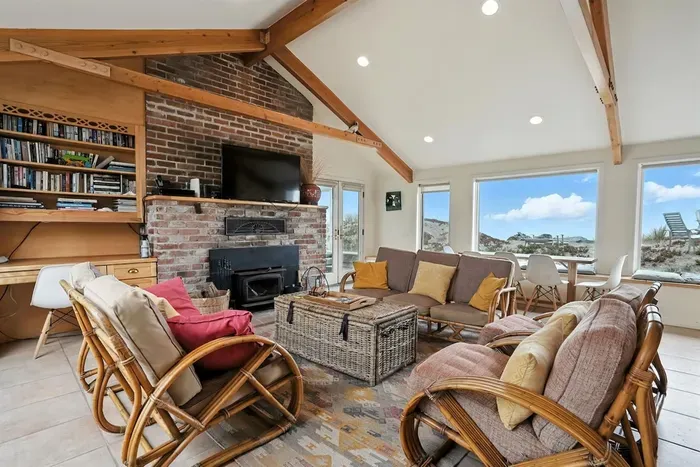 - Cozy living room with vaulted ceilings, rattan furniture, and vibrant pillows  - Brick fireplace with mounted TV for a warm and inviting atmosphere  - Large windows offer sweeping views of the dunes and ocean beyond