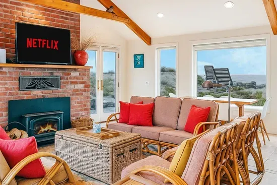 - Cozy living room with rattan seating, vibrant cushions, and brick fireplace  - Large windows frame beautiful ocean views for a relaxing seaside vibe  - Wall mounted TV with streaming options ensures entertainment for all guests