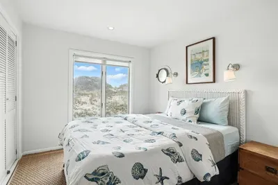 - Light filled bedroom with coastal bedding and sliding glass door to outdoors  - Features porthole style window, nightstands, and wall mounted lights  - Serene setting with soft tones and dune views from the bed