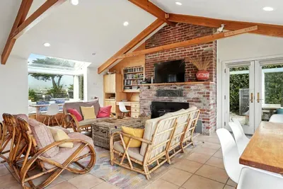 - Spacious open living area with multiple seating zones and large dining table  - Brick fireplace and exposed beams enhance the cozy yet airy design  - Walls of windows showcase stunning outdoor views from every angle