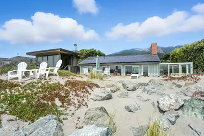 Beachfront home with outdoor seating surrounded by sand and rocks.