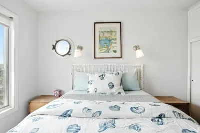 - Bright coastal themed bedroom with white headboard and sea inspired bedding  - Features porthole style window, wall sconces, and framed art  - Crisp white walls and light wood furniture enhance the airy atmosphere