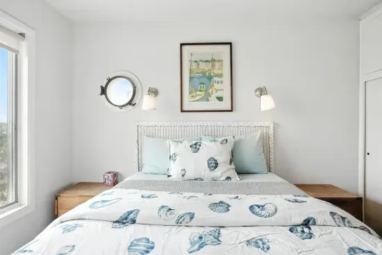 - Bright coastal themed bedroom with white headboard and sea inspired bedding  - Features porthole style window, wall sconces, and framed art  - Crisp white walls and light wood furniture enhance the airy atmosphere