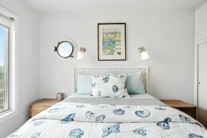 - Bright coastal themed bedroom with white headboard and sea inspired bedding  - Features porthole style window, wall sconces, and framed art  - Crisp white walls and light wood furniture enhance the airy atmosphere