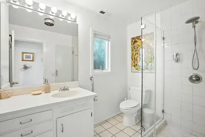 - Modern white bathroom with glass enclosed shower and tile flooring  - Features vanity with large mirror, bright lighting, and plenty of counter space  - Window allows soft daylight for a fresh, airy ambiance
