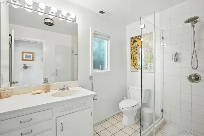 - Modern white bathroom with glass enclosed shower and tile flooring  - Features vanity with large mirror, bright lighting, and plenty of counter space  - Window allows soft daylight for a fresh, airy ambiance