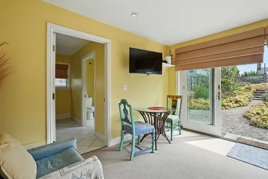 - Bright sitting area with French doors opening to the garden and patio seating  - Includes a small dining set, wall mounted TV, and easy access to the bathroom  - Cheerful yellow walls create a warm and inviting space for guests to enjoy