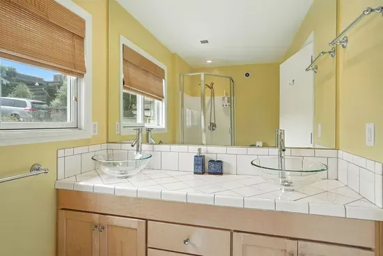 - Bright bathroom with dual glass vessel sinks and large mirror  - Tiled countertop and natural wood cabinetry create a warm, inviting feel  - Walk in shower and windows bring in natural light for a fresh start to the day