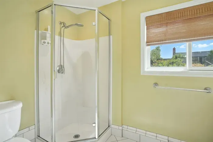 - Bright bathroom with corner glass shower and sleek fixtures for a modern touch  - Window with bamboo shade allows soft daylight while maintaining privacy  - Light yellow walls create a cheerful and inviting atmosphere