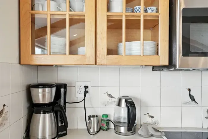 - Coffee station with drip coffee maker, electric kettle, & essential supplies for your morning  - Glass front cabinets neatly display cups, plates, & bowls for easy access  - Decorative tile accents with coastal bird designs add charm to the space