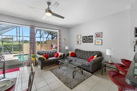 - Bright living room with plush gray sofas, vibrant red cushions, and wall art  
- Sliding doors open to the pool area for easy indoor outdoor flow and relaxation  
- Ceiling fan and natural light create a fresh, welcoming atmosphere year round
