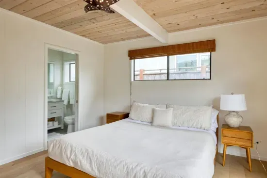 - Queen bedroom with crisp white bedding and warm wood furniture accents  
- Large window above the bed lets in light to brighten the cozy retreat  
- Convenient en suite bath access provides privacy during your stay
