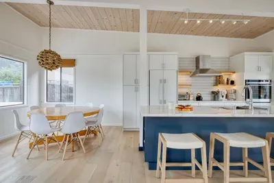- Dining space with round table and modern chairs for shared meals  
- Kitchen features a navy island with bar seating for casual dining  
- Light wood flooring and ceiling create a warm, coastal inspired vibe