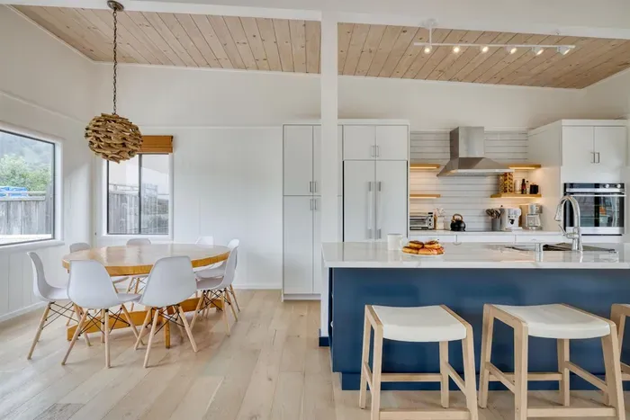 - Dining space with round table and modern chairs for shared meals  
- Kitchen features a navy island with bar seating for casual dining  
- Light wood flooring and ceiling create a warm, coastal inspired vibe