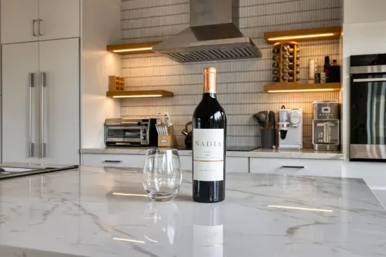 - Elegant kitchen island with marble countertop sets a refined tone  
- Bottle of wine and glass ready to welcome guests upon arrival  
- Modern backsplash, open shelving, and under cabinet lighting add flair