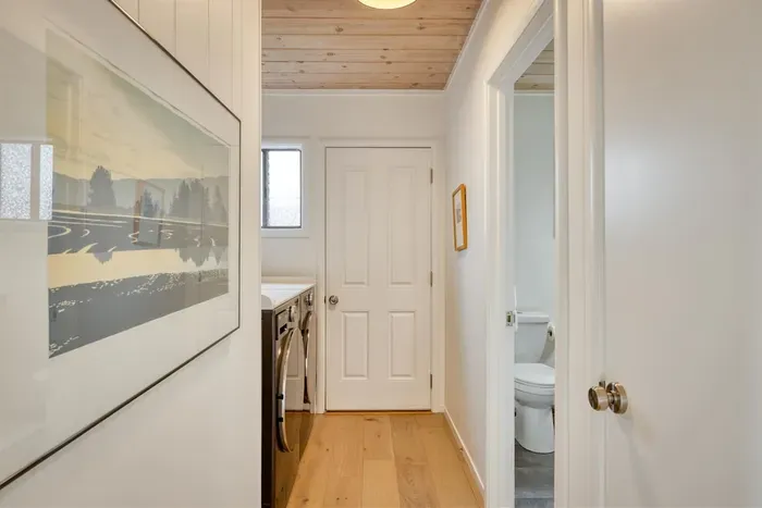 - Hallway with warm wood flooring and ceiling leading to laundry and bath  
- Side window allows natural light to brighten the functional corridor  
- Minimal wall art adds charm to this simple yet practical connecting space