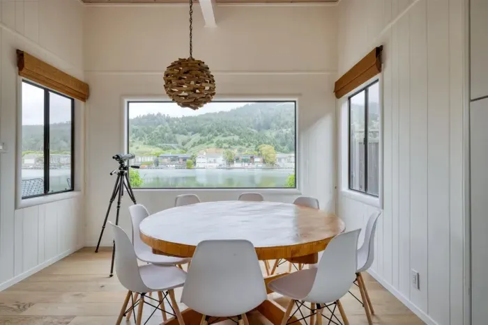 - Bright dining area with round wooden table and seating for eight guests  
- Stylish windows offer panoramic water and hillside views while dining  
- Unique pendant light adds charm to this airy, scenic gathering space