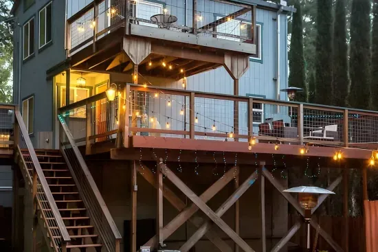 - Elevated two story home with wraparound deck and string lights for charm  
- Outdoor seating with fire pit creates a cozy gathering space at night  
- Surrounded by tall pines for a peaceful and private retreat experience