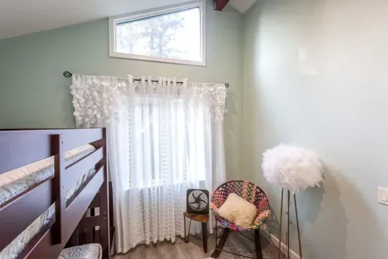 - Cozy bedroom corner with wooden bunk beds and colorful accent chair  
- Soft natural light through sheer curtains creates a gentle atmosphere  
- High ceiling with transom window adds openness to the intimate space