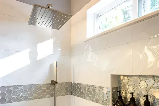 - Sleek shower with rainfall head, handheld sprayer & contemporary hardware finishes
- White textured tiles accented with metallic hexagonal band for a designer look
- Built in recessed shelf keeps bath products organized & within easy reach for use