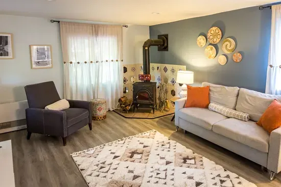 - Welcoming living room with wood burning stove framed by decorative tile surround  
- Plush sofa & accent pillows create a warm & inviting spot to unwind & relax  
- Sunlight filters through soft curtains, adding to the room’s cozy charm & appeal