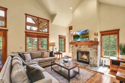 The open concept main floor at Alpine Escape features a cozy living area with a warm wood fireplace and 24-foot vaulted ceilings, perfect for relaxing after a day on the slopes.