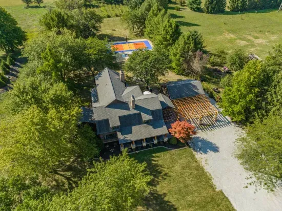 Aerial view of a spacious countryside home surrounded by lush greenery, featuring a large driveway, covered patio area, and a private outdoor basketball court in the backyard.
