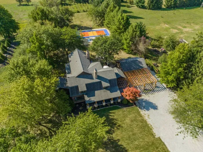 Aerial view of a spacious countryside home surrounded by lush greenery, featuring a large driveway, covered patio area, and a private outdoor basketball court in the backyard.