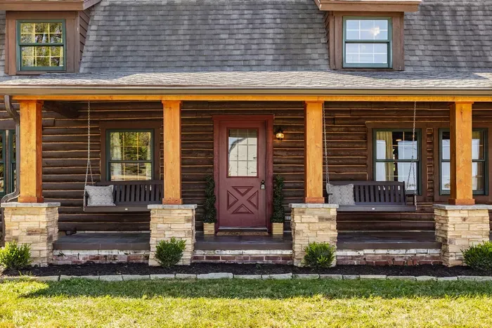 Inviting entryway featuring log-style architecture with stone pillars and matching wood accents. Two hanging benches frame the red front door, blending craftsmanship and comfort in a timeless rustic design.