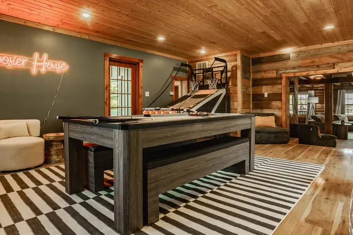 Game room with wood accents, a pool table, basketball arcade game, and lounge area under a glowing “Hoosier House” sign.