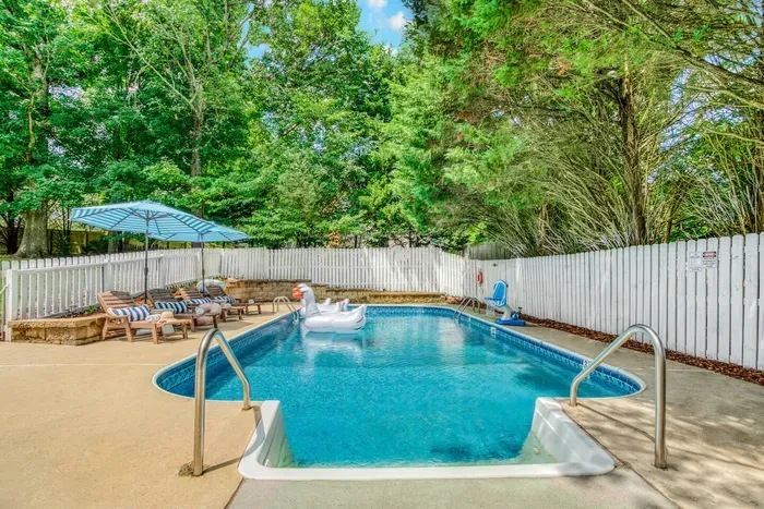 Private pool surrounded by lush greenery and a white fence, featuring lounge chairs with striped cushions and umbrellas