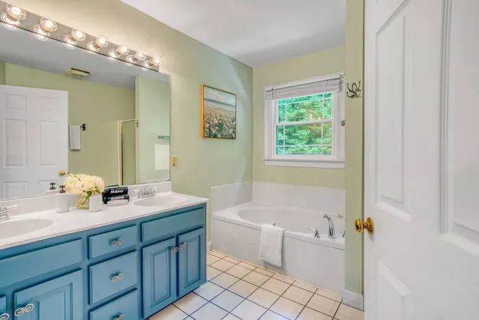 A bright and airy bathroom featuring a deep soaking tub, double-sink vanity with stylish blue cabinets, and a large mirror—perfect for unwinding or getting ready in comfort.