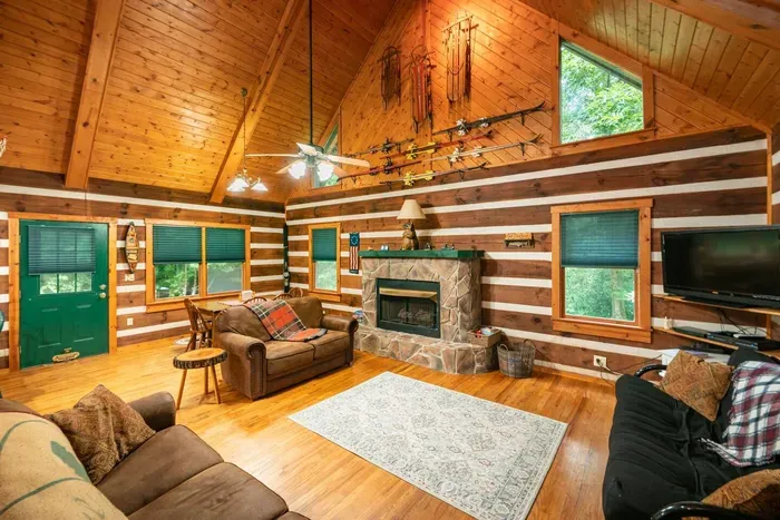 Warm and inviting log cabin living room featuring vaulted wood ceilings, a cozy stone fireplace, comfortable seating, rustic décor, and large windows that fill the space with natural light.