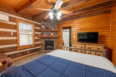 Cozy rustic cabin bedroom featuring a comfortable bed with blue bedding, warm wood interiors, a stone fireplace, flat-screen TV, ample storage, and windows bringing in plenty of natural light.