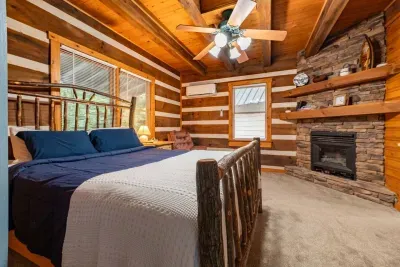 Cozy rustic cabin bedroom featuring a comfortable bed with blue bedding, warm wood interiors, a stone fireplace, flat-screen TV, ample storage, and windows bringing in plenty of natural light.