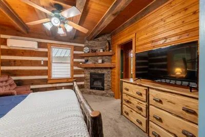 Cozy rustic cabin bedroom featuring a comfortable bed with blue bedding, warm wood interiors, a stone fireplace, flat-screen TV, ample storage, and windows bringing in plenty of natural light.