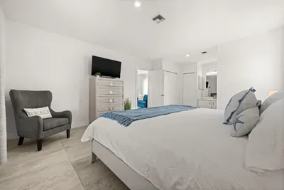 Bright coastal-style bedroom with TV, lounge chair, and en suite access
