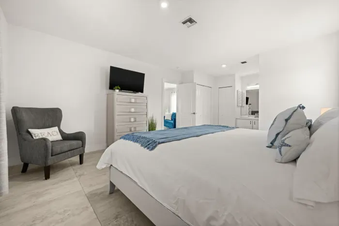 Bright coastal-style bedroom with TV, lounge chair, and en suite access