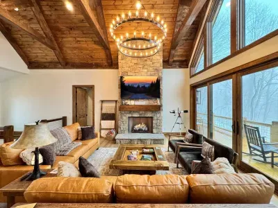Spacious family room with plush leather sofas, a 75" smart TV, and a cozy fireplace. Enjoy breathtaking mountain views through the window wall with sliding glass doors, offering a perfect blend of comfort and natural beauty.