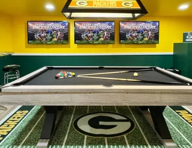 The downstairs Packer game room has a pool table, 3 55" TVs, 2 Packer pub tables with stools, a leather couch, and Golden Tee golf video game.
