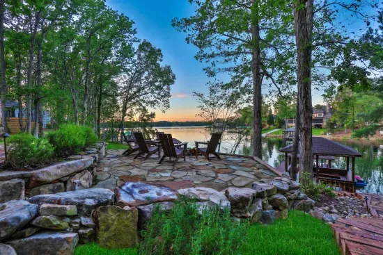 A peaceful stone patio overlooks the calm lake, framed by mature trees and Adirondack chairs and firepit — an ideal spot to sip coffee at sunrise or unwind with a glass of wine at sunset.