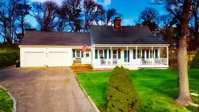 A charming white Cape Cod-style home featuring a spacious front porch with comfortable chairs and a three-car driveway parking.