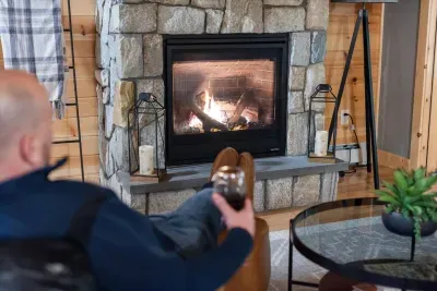 A cozy fire crackles softly, casting a warm glow on the room. Feet rest comfortably on an ottoman, a glass of wine in hand, as the soothing flames create a perfect setting for relaxation. 