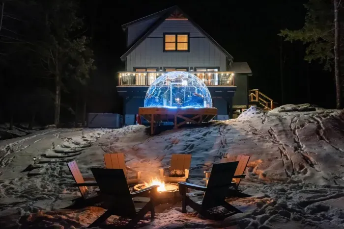 A serene nighttime backyard with golden light pouring from a sleek luxury dome. A glowing fire pit crackles nearby, casting shadows over the steaming hot tub. Warm ambient lighting wraps the scene, creating a cozy, intimate escape under the stars.