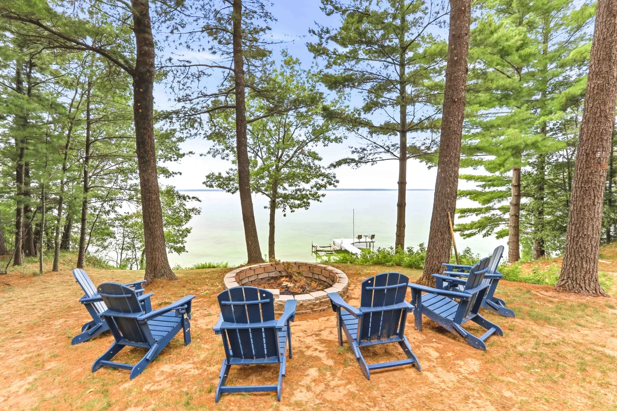 Brick fire pit with six blue Adirondack chairs on sand by a lake with pine trees, at Ashley's Hideaway, a Clear Trail