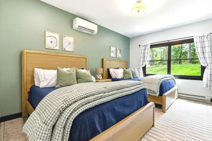 Rest easy in a bright double-bedroom retreat with layered linens, warm wood tones, and peaceful green views just outside the window.