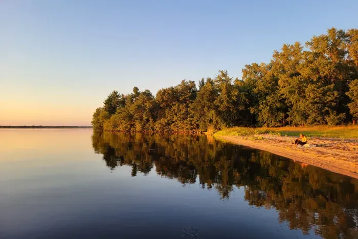 Unwind on the quiet sandy shore as golden-hour light reflects across calm water and the tree-lined coastline.