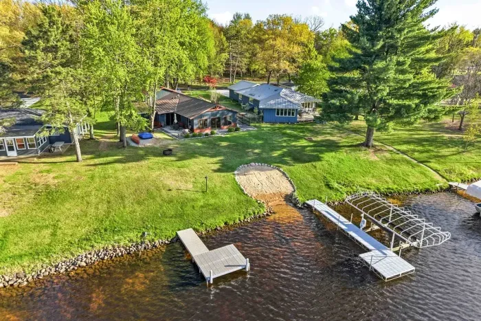 Beautiful aerial lakefront view showcasing a sandy shoreline, private dock access, and a spacious green yard surrounded by mature trees.