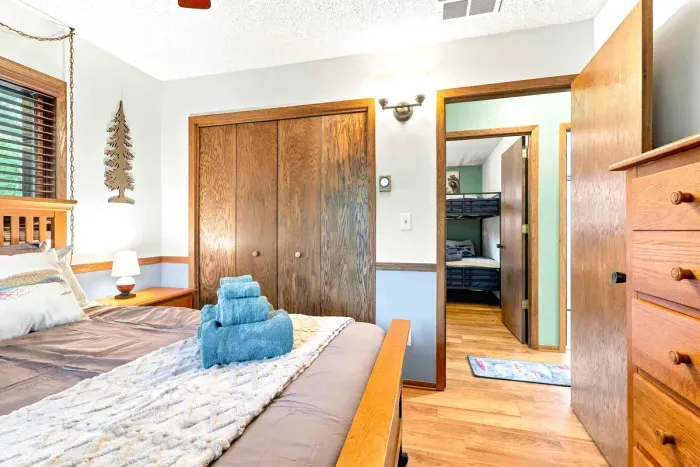 Relax in a bright, peaceful bedroom styled with classic wood accents, a comfortable wooden bed, and a convenient nightstand. Wake up to sunlight streaming through the windows framed by tranquil forest scenery.